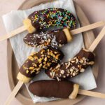 Overhead view of a platter lined with parchment with five chocolate covered banana popsicles with various toppings.