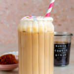 A glass of iced sugar cookie latte topped with sugar cookie sweet cream and sprinkles with a straw. Sugar cookie syrup in the background with sprinkles scattered around and brown sugar on the side.