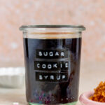 A glass jar of sugar cookie syrup, labeled. Sprinkles in front with some brown sugar on the side.