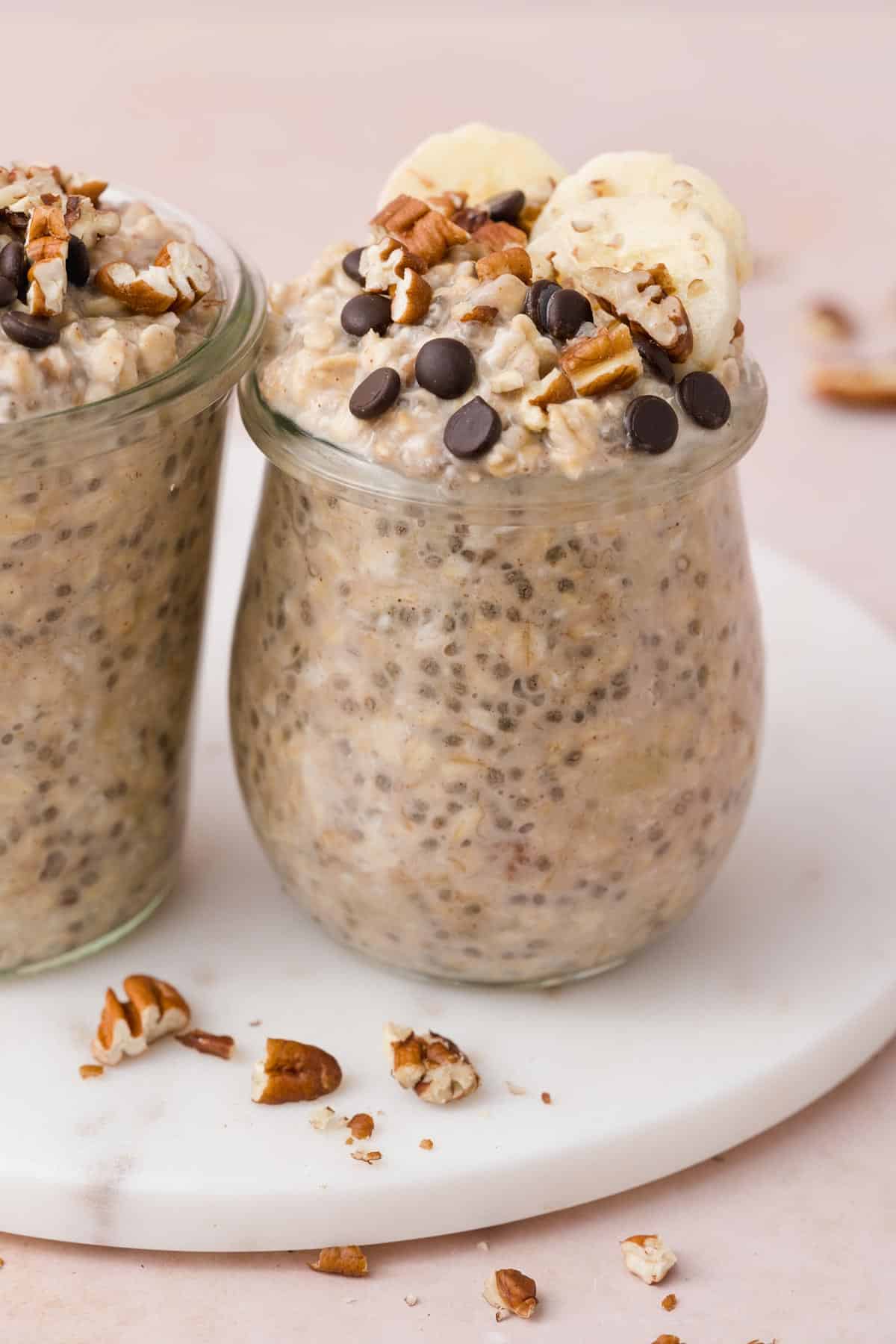 A jar of banana bread overnight oats topped with chocolate chips, chopped pecans, and banana slices on a marble slab. A second jar off to the side.