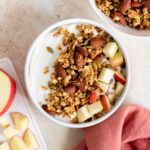 A bowl of yogurt topped with applesauce granola and diced apples.