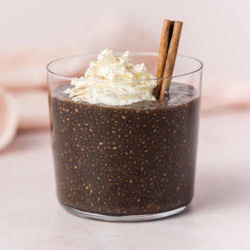 A glass of gingerbread chia pudding topped with whipped cream, a cinnamon stick, and a dusting of cinnamon powder.