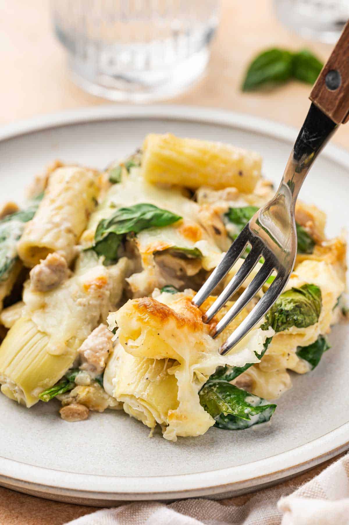 A fork pressed into a bite of spinach and artichoke pasta bake on a plate.
