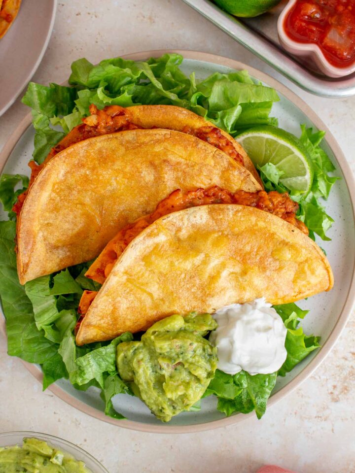 Two crispy baked ground chicken tacos over lettuce with a dollop of guacamole, sour cream, and a lime on a plate.