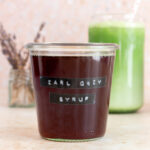 A glass of earl grey syrup with an iced matcha in the background with dried lavender in a small vase.