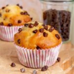 A greek yogurt chocolate chip muffin on a parchment-lined serving board with another muffin in the back along with a glass of mini chocolate chips.