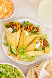 An overhead view of a plate with three ground turkey tacos with sour cream, guacamole, shredded cheese, and lettuce. Lime wedges on the plate.