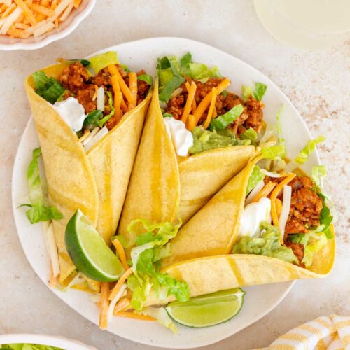 An overhead view of a plate with three ground turkey tacos with sour cream, guacamole, shredded cheese, and lettuce. Lime wedges on the plate.