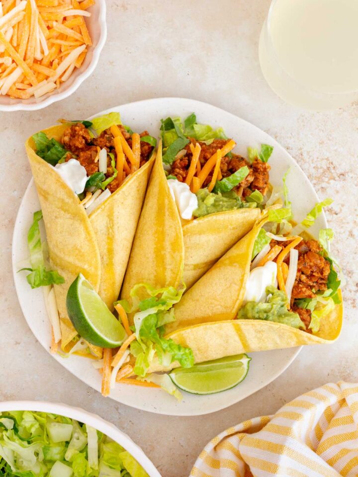 An overhead view of a plate with three ground turkey tacos with sour cream, guacamole, shredded cheese, and lettuce. Lime wedges on the plate.