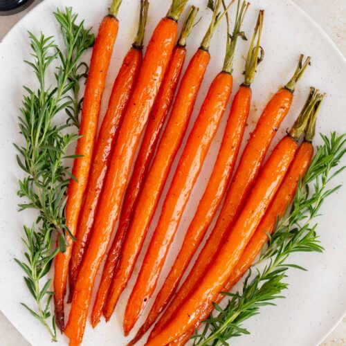 A white platter of maple glazed carrots with some herbs surrounding it.
