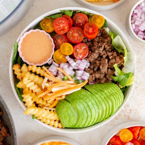 Overhead view of a burger bowl (avocado, ground beef, tomatoes, fries, red onions, and shredded cheese over lettuce) with a bowl of special sauce.