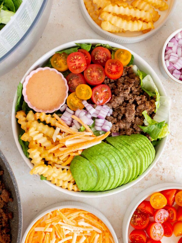 Overhead view of a burger bowl (avocado, ground beef, tomatoes, fries, red onions, and shredded cheese over lettuce) with a bowl of special sauce.