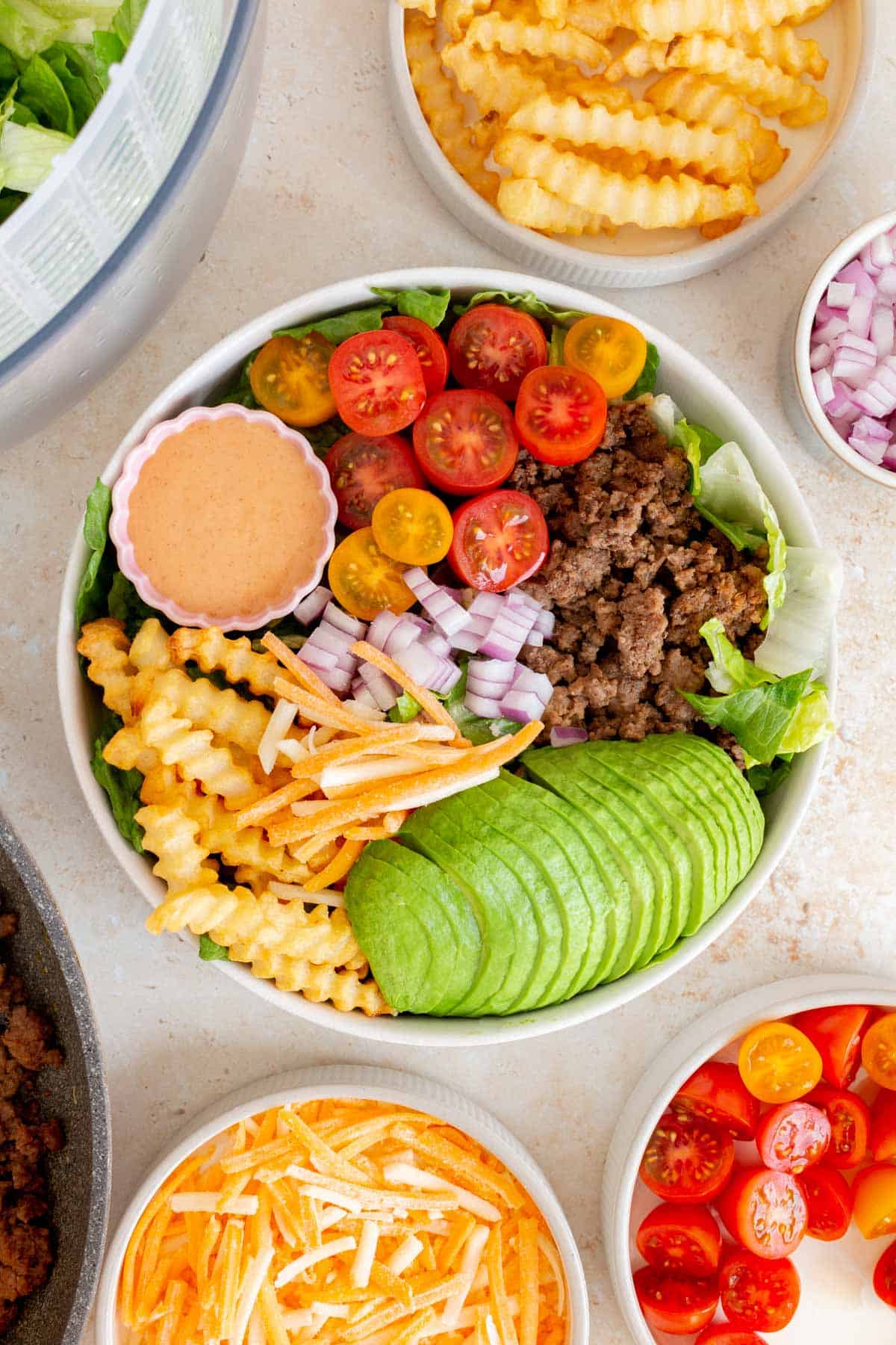 Overhead view of a burger bowl (avocado, ground beef, tomatoes, fries, red onions, and shredded cheese over lettuce) with a bowl of special sauce.