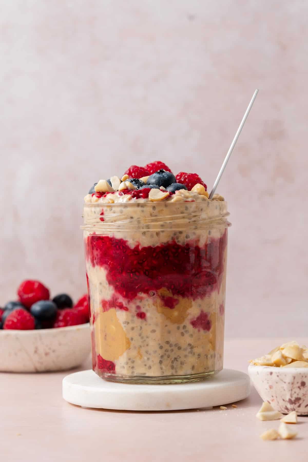 A profile view of a jar of peanut butter and jelly overnight oats on a coaster topped with fresh berries and peanuts.