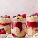 A profile view of a jar of peanut butter and jelly overnight oats on a coaster topped with fresh berries and peanuts. More jars of overnight oats in the background.