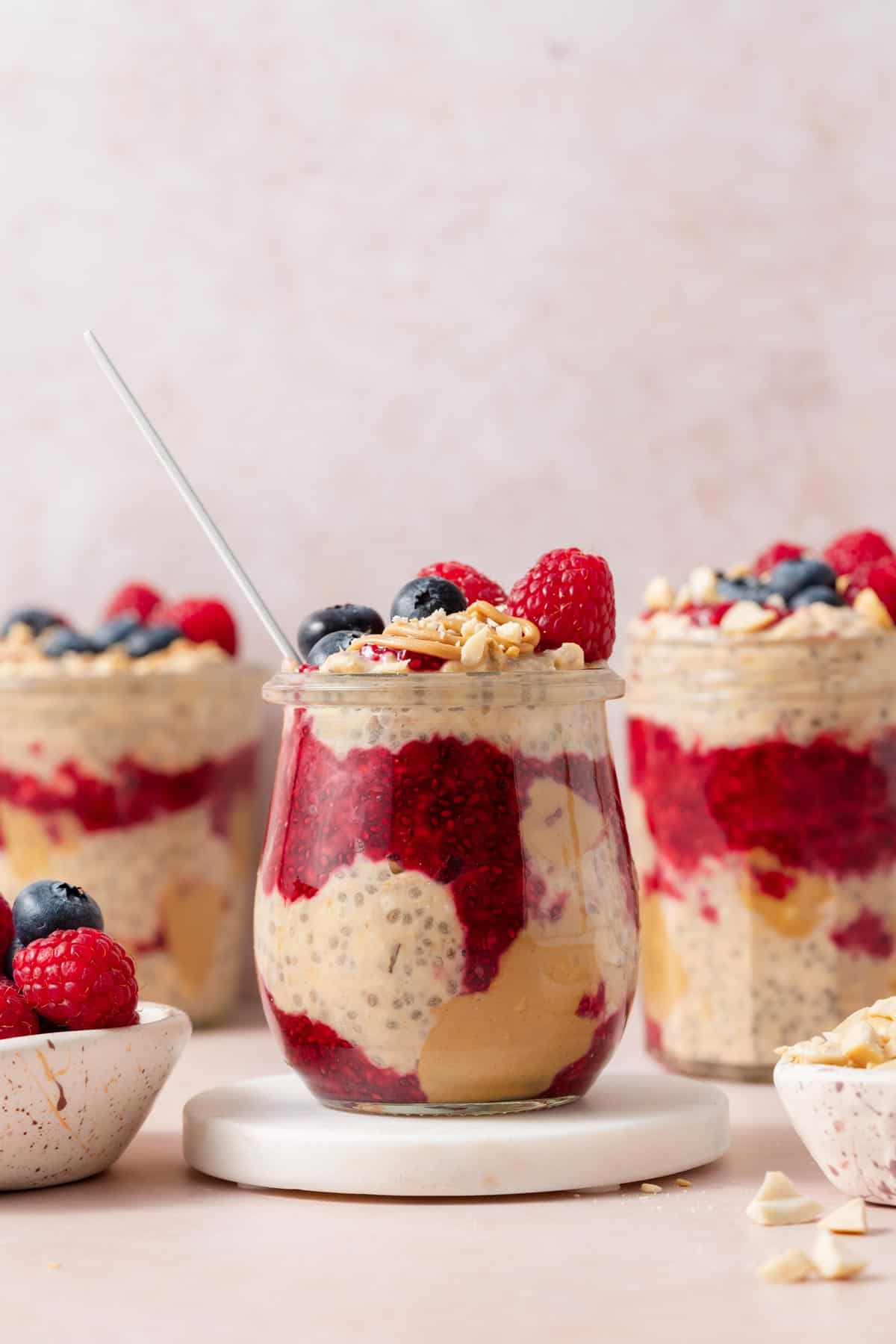 A profile view of a jar of peanut butter and jelly overnight oats on a coaster topped with fresh berries and peanuts. More jars of overnight oats in the background.