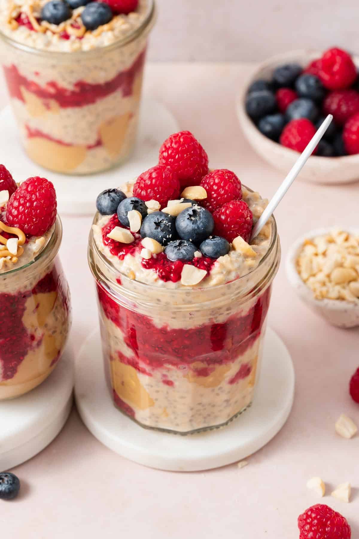 A slightly overhead view of a jar of peanut butter and jelly overnight oats on a coaster, topped with peanuts, blueberries, and raspberries.