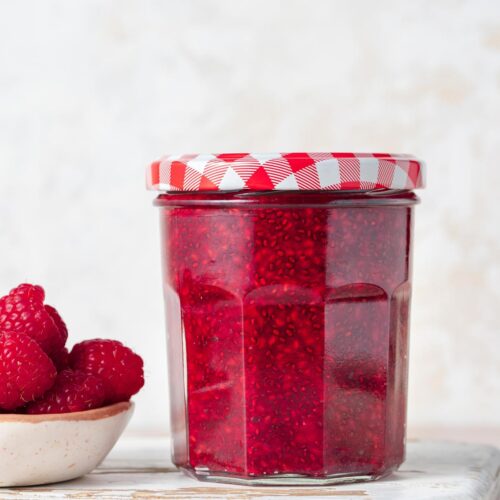 A profile view of a jar of homemade raspberry chia jam. A small bowl of fresh raspberries on the side and one berry on the other side.