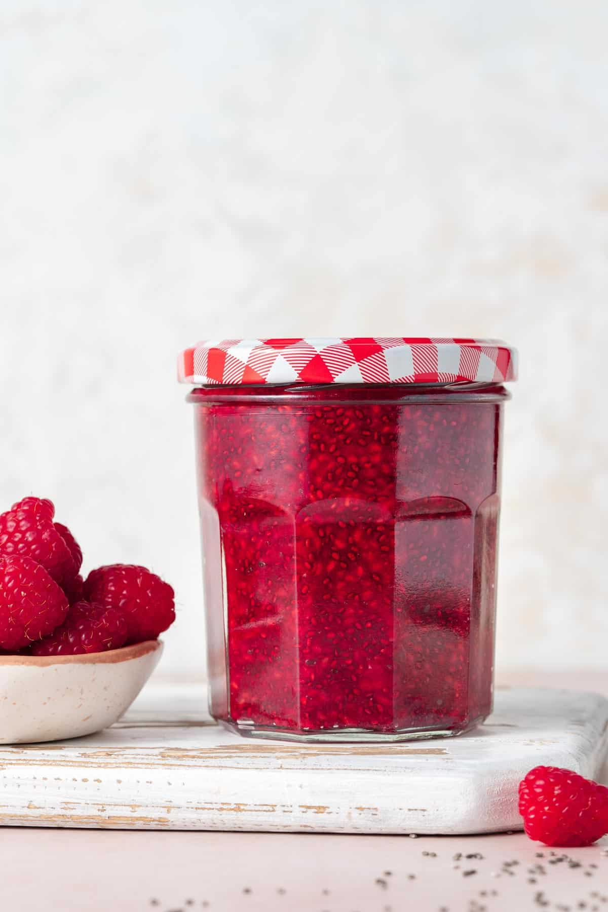 A profile view of a jar of homemade raspberry chia jam. A small bowl of fresh raspberries on the side and one berry on the other side.