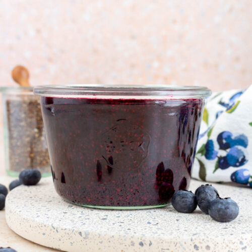 A jar of blueberry chia jam with fresh blueberries scattered around.