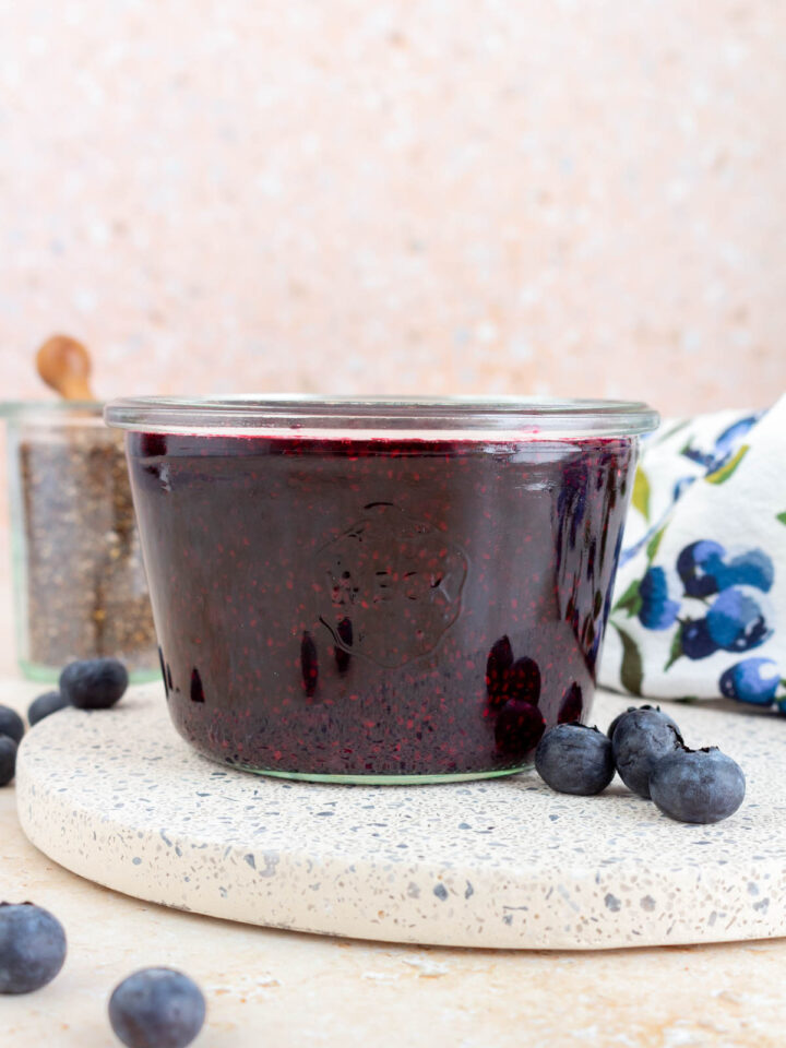 A jar of blueberry chia jam with fresh blueberries scattered around.