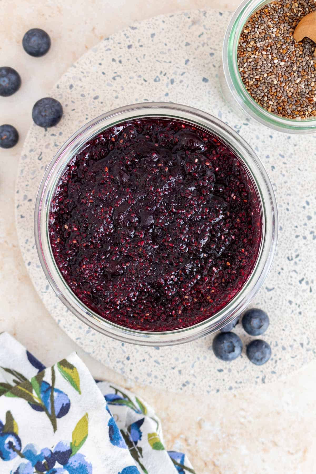 Overhead view of a jar of blueberry chia jam with fresh blueberries scattered around and a jar of chia seeds.
