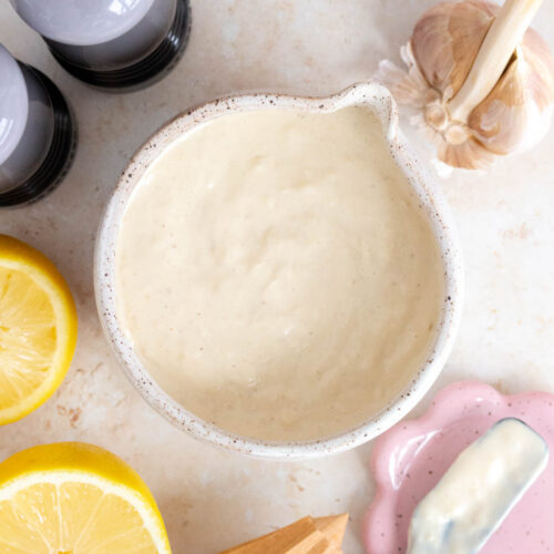 Overhead view of a bowl of homemade caesar salad dressing. Cut lemon, garlic, salt and pepper shaker, and a lemon juicer on the side.