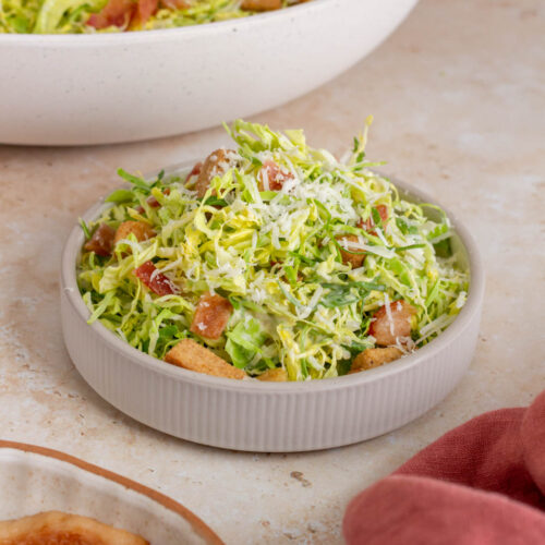A small bowl of shaved brussels sprouts caesar salad piled high. More salad in the background.