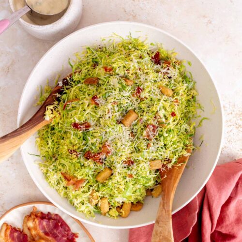 Overhead view of a large bowl of shaved brussels sprouts caesar salad with wooden serving utensils. Some bacon on the side and more dressing on the side.