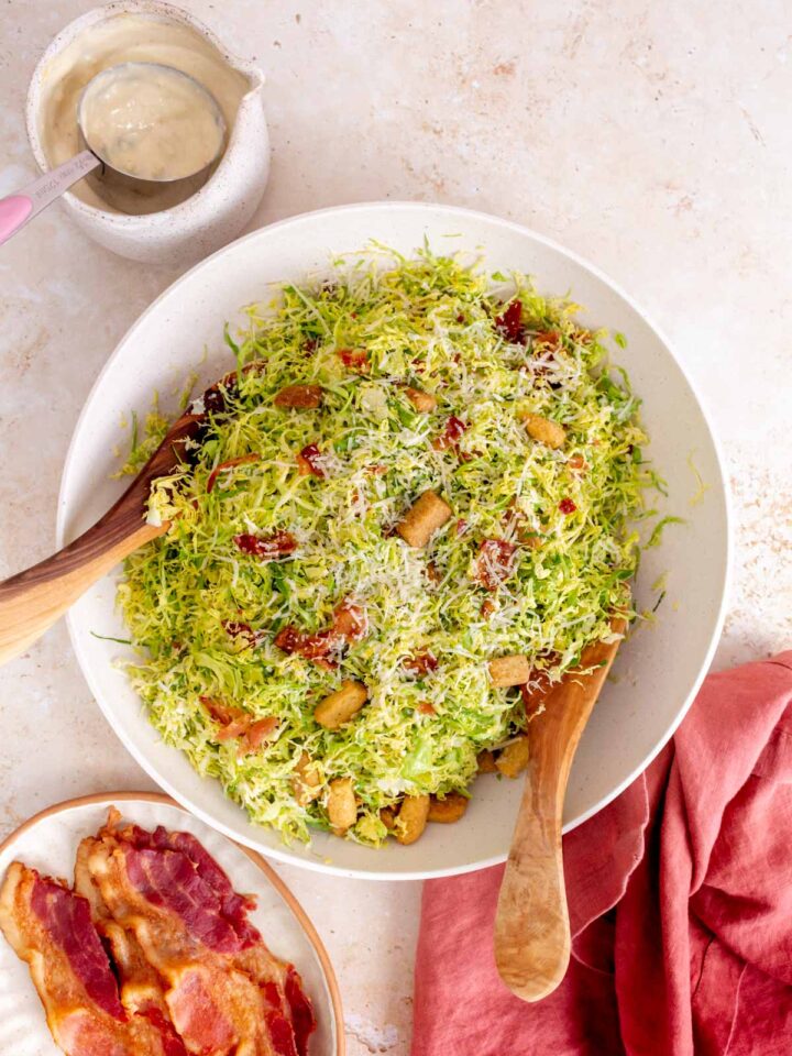 Overhead view of a large bowl of shaved brussels sprouts caesar salad with wooden serving utensils. Some bacon on the side and more dressing on the side.