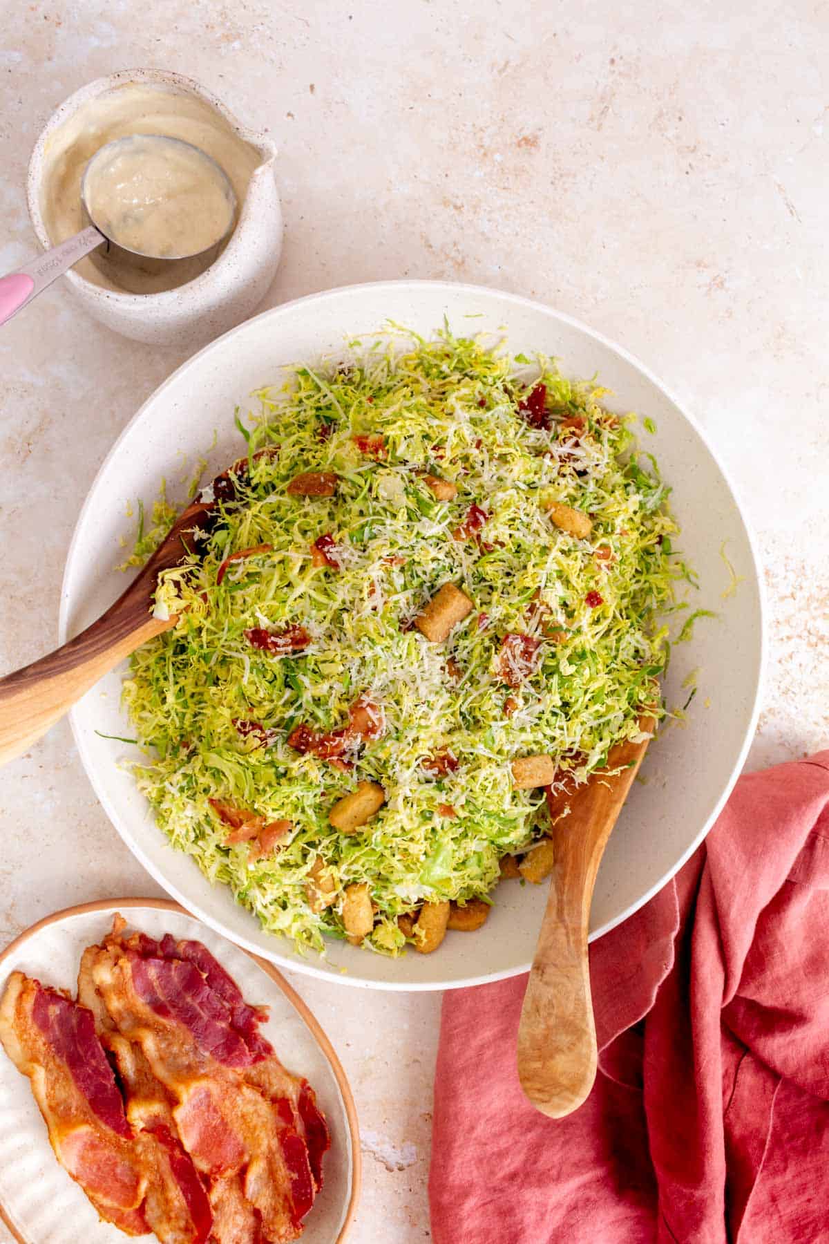 Overhead view of a large bowl of shaved brussels sprouts caesar salad with wooden serving utensils. Some bacon on the side and more dressing on the side.