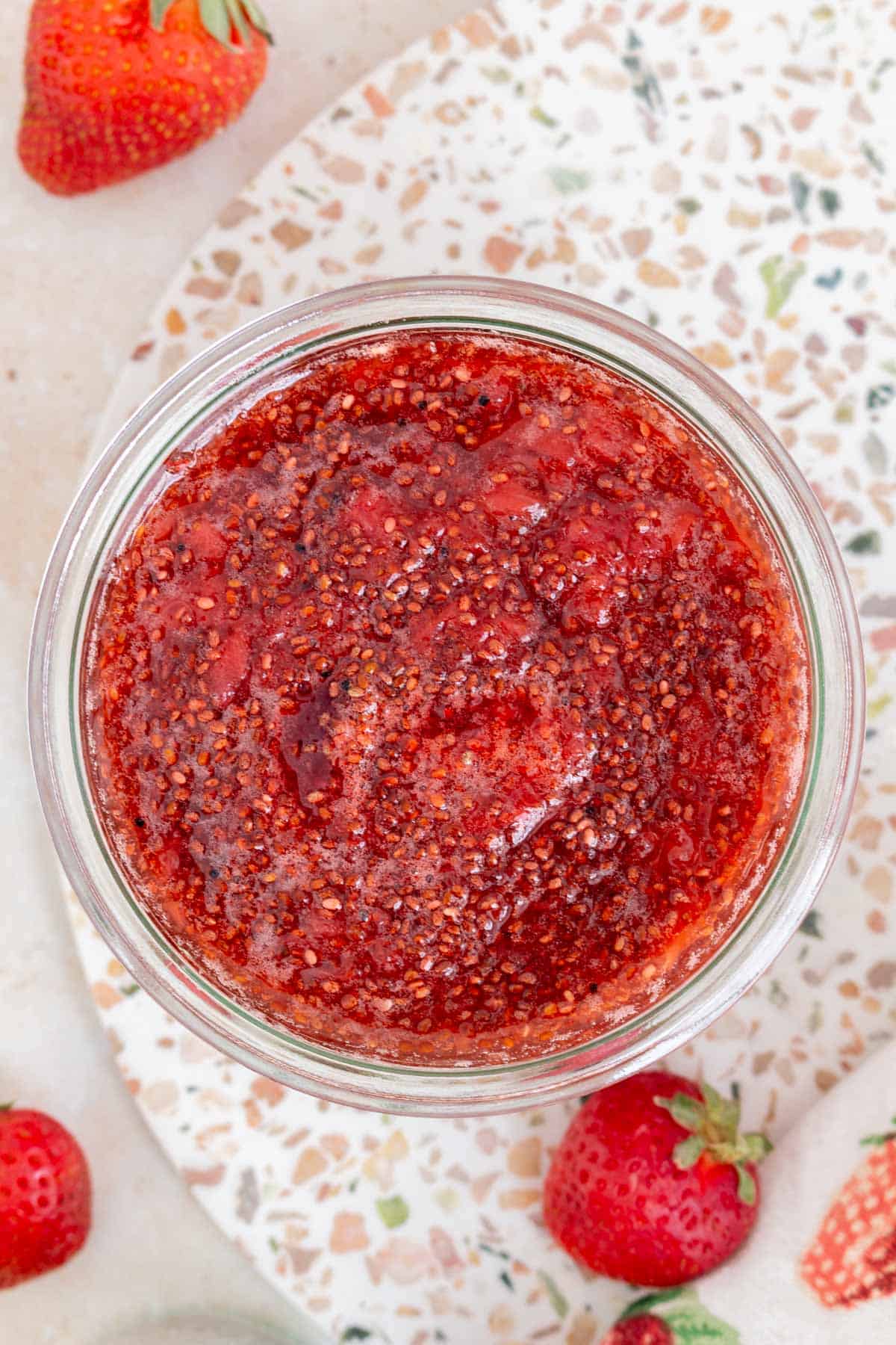 Overhead view of a jar of strawberry chia jam. Fresh strawberries scattered around.