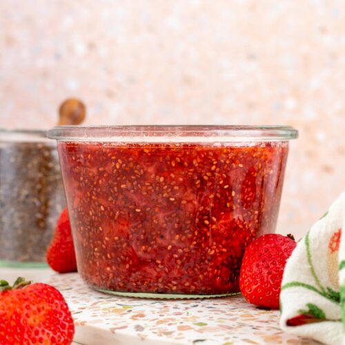 A profile view of a jar of strawberry chia jam. Fresh strawberry scattered around with a jar of chia seeds in the background.