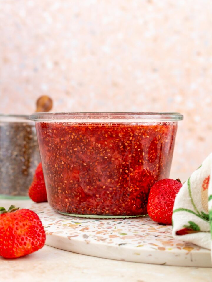 A profile view of a jar of strawberry chia jam. Fresh strawberry scattered around with a jar of chia seeds in the background.