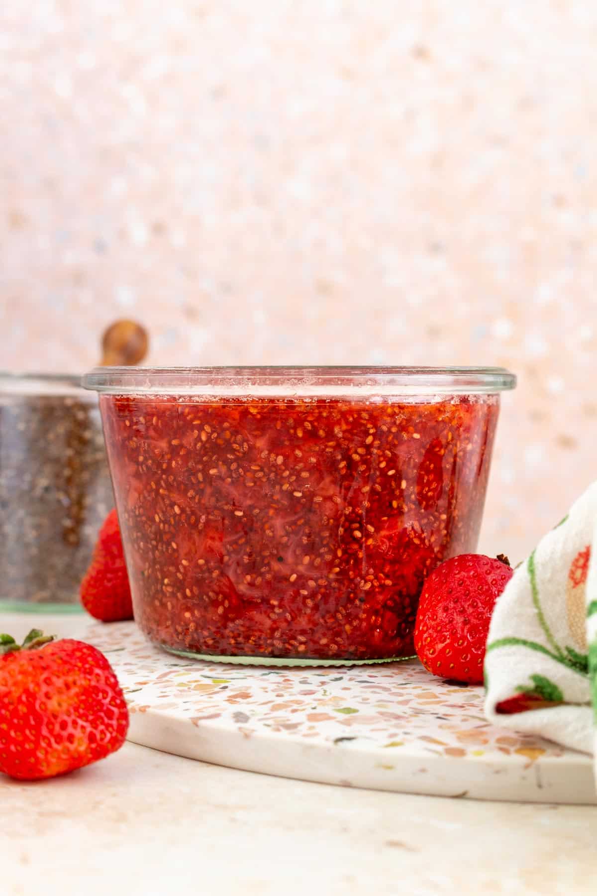 A profile view of a jar of strawberry chia jam. Fresh strawberry scattered around with a jar of chia seeds in the background.