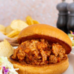 A plate with turkey sloppy joes on a broiche bun. Coleslaw and chips on the side.