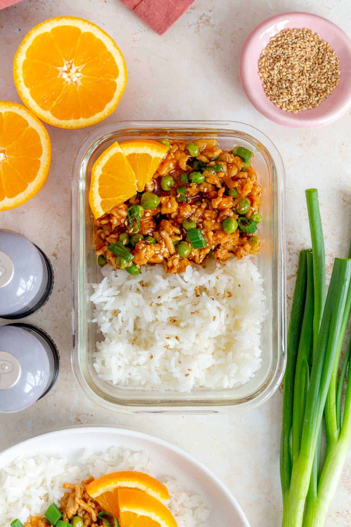 A meal prep container of volcano chicken with rice topped with sesame seeds, green onions, and orange slices.