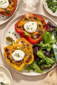 A plate with two taco stuffed peppers with extra veggies topped with sour cream on a plate along with some salad.