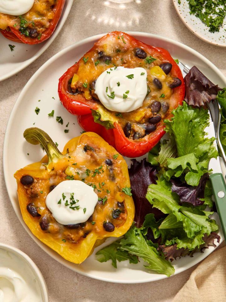 A plate with two taco stuffed peppers with extra veggies topped with sour cream on a plate along with some salad.