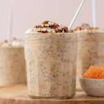 A profile view of a jar of carrot cake overnight oats topped with chopped pecans with a bowl of shredded carrots. More jars in the background.