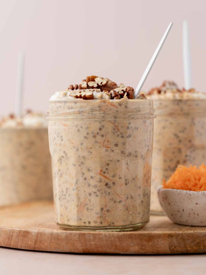 A profile view of a jar of carrot cake overnight oats topped with chopped pecans with a bowl of shredded carrots. More jars in the background.