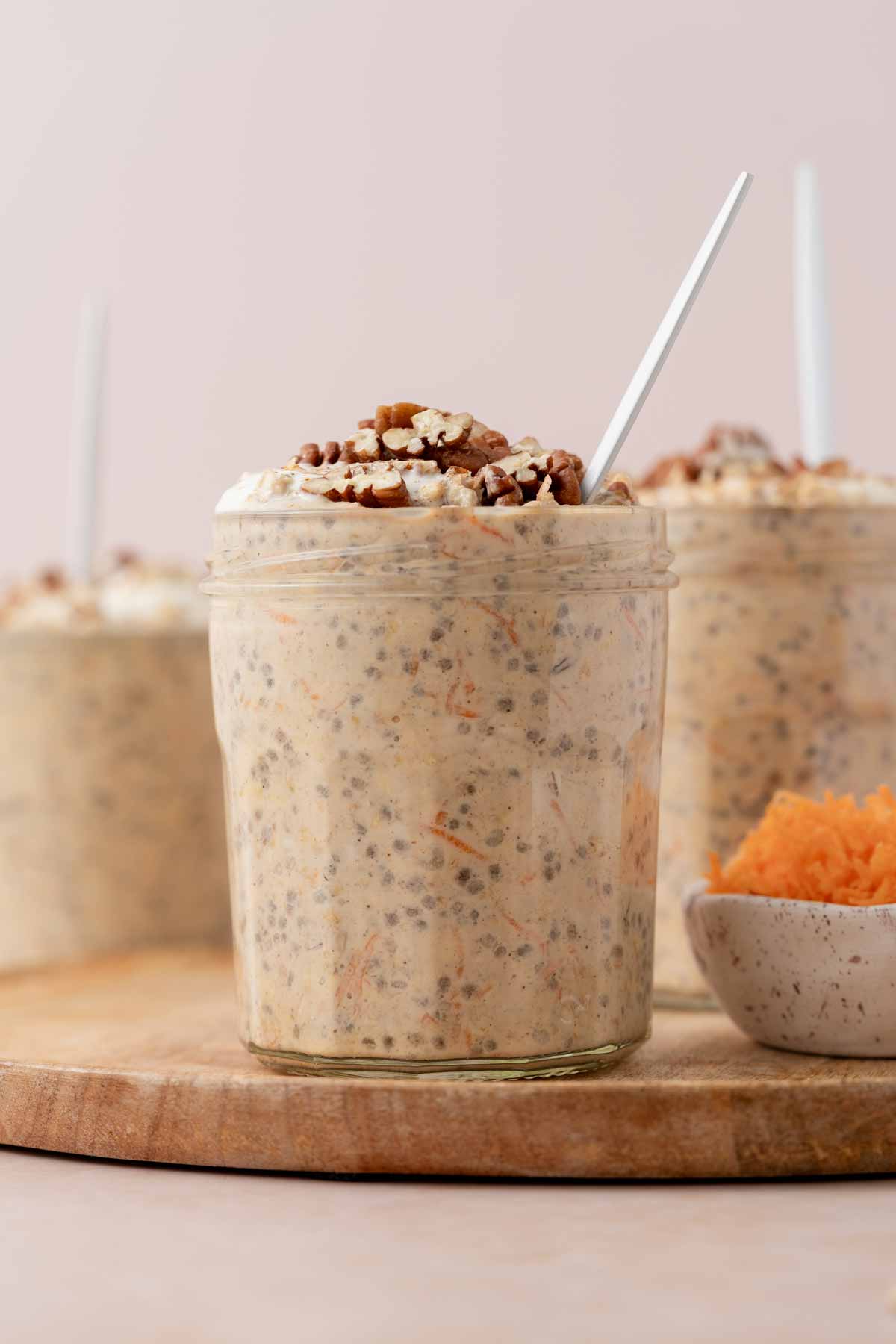 A profile view of a jar of carrot cake overnight oats topped with chopped pecans with a bowl of shredded carrots. More jars in the background.