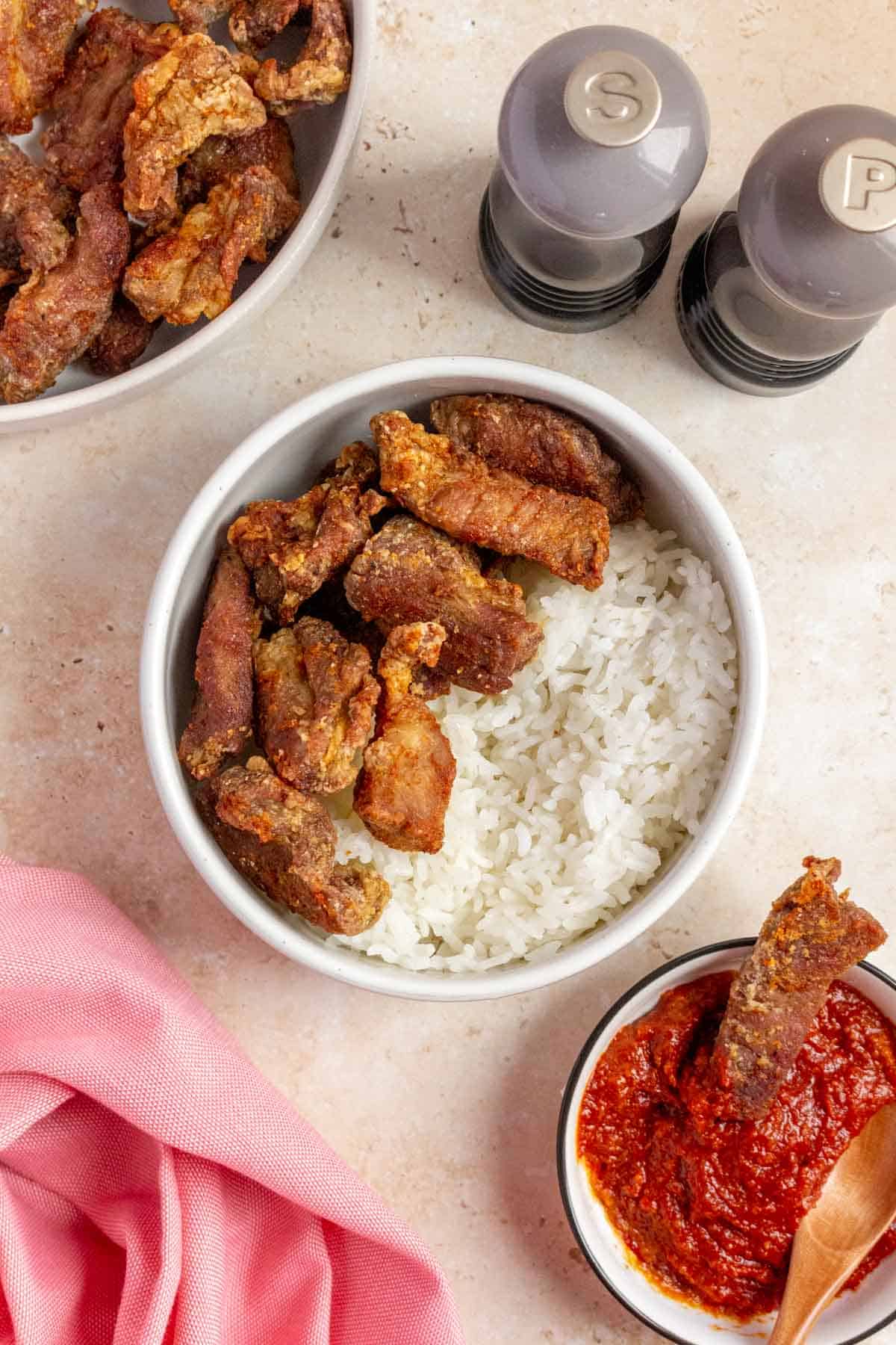 A bowl with rice and air fryer salt and pepper pork bites. Salt and pepper shakers on the side along with dipping sauce with one piece of pork.