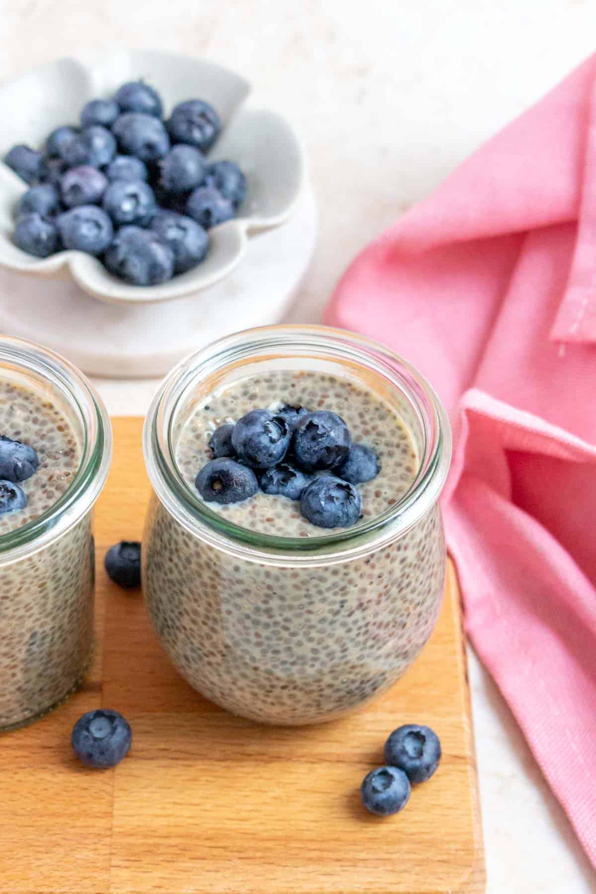 A slightly overhead view of a jar of earl grey chia pudding topped with blueberries on a wooden serving board. Another jar off to the side and a bowl of blueberries in the background.
