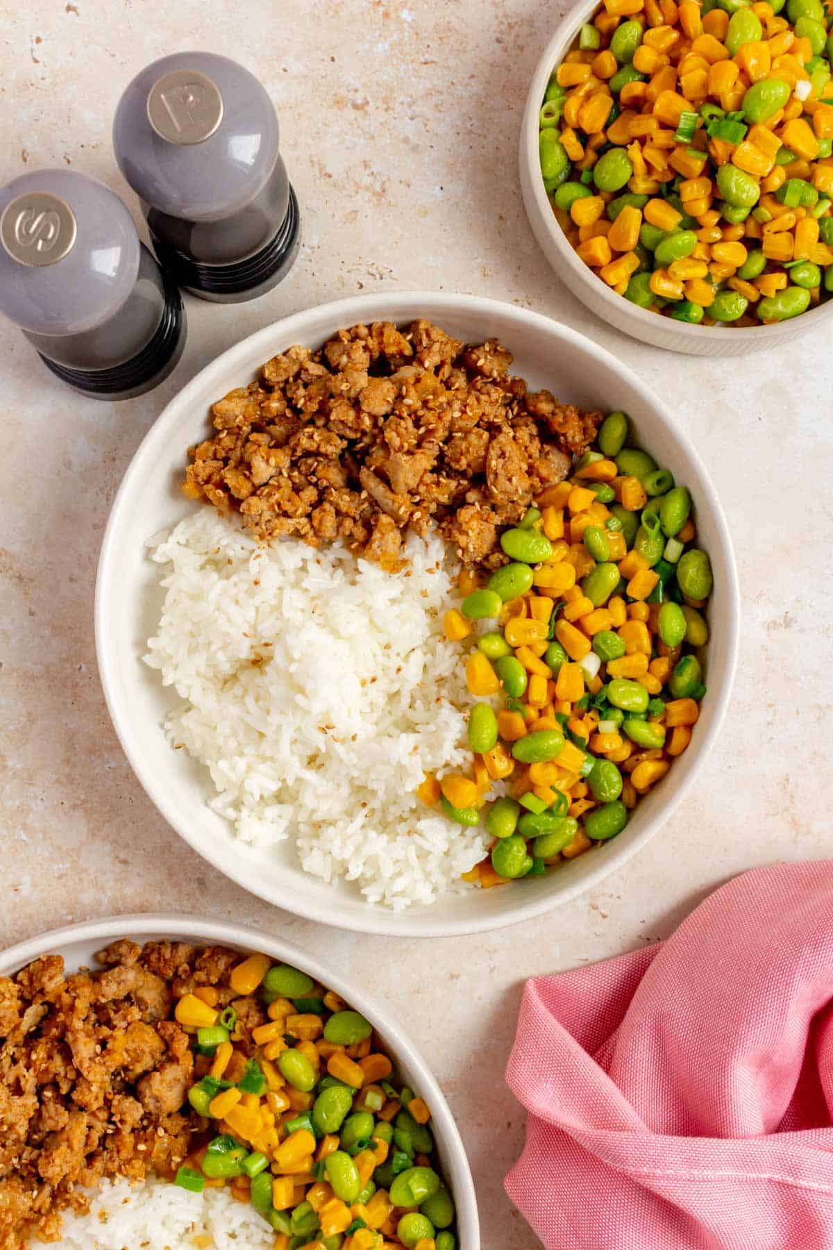 An overhead view of a hot honey ground pork bowl with corn and edamame. Salt and pepper on the side along with another bowl and a plate of just corn and edamame.