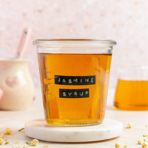 A weck jar of jasmine simple syrup with a label saying "jasmine syrup" on a round marble coaster. Jasmine flowers scattered around.