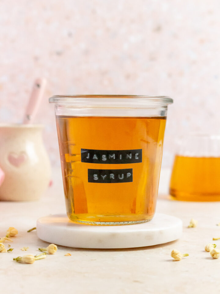 A weck jar of jasmine simple syrup with a label saying "jasmine syrup" on a round marble coaster. Jasmine flowers scattered around.