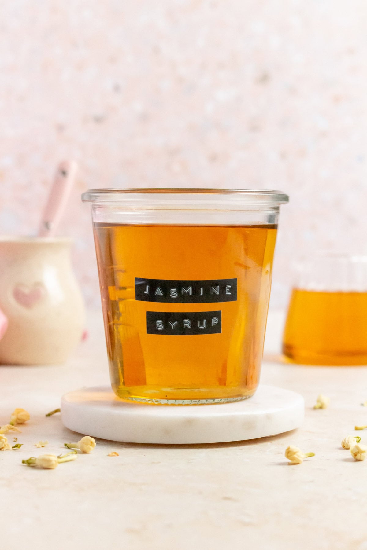 A weck jar of jasmine simple syrup with a label saying "jasmine syrup" on a round marble coaster. Jasmine flowers scattered around.