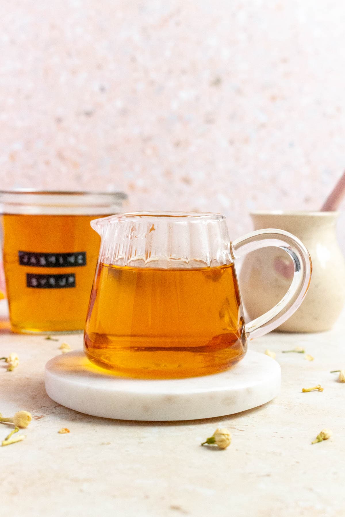 A small jug of jasmine syrup on a marble coaster. Dried jasmine flowers scattered around the surface.