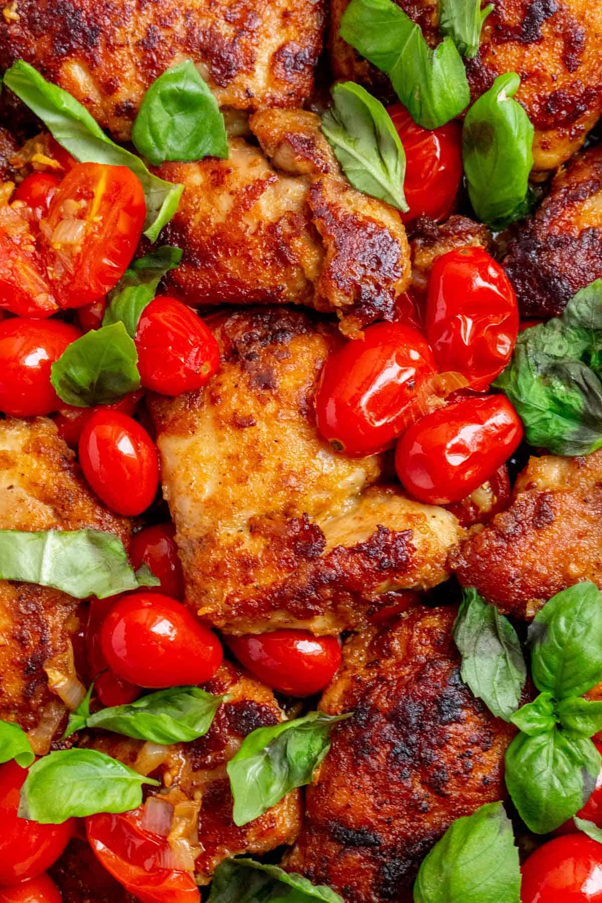 Close up view of crispy chicken thighs surrounded by grape tomatoes and fresh basil leaves.